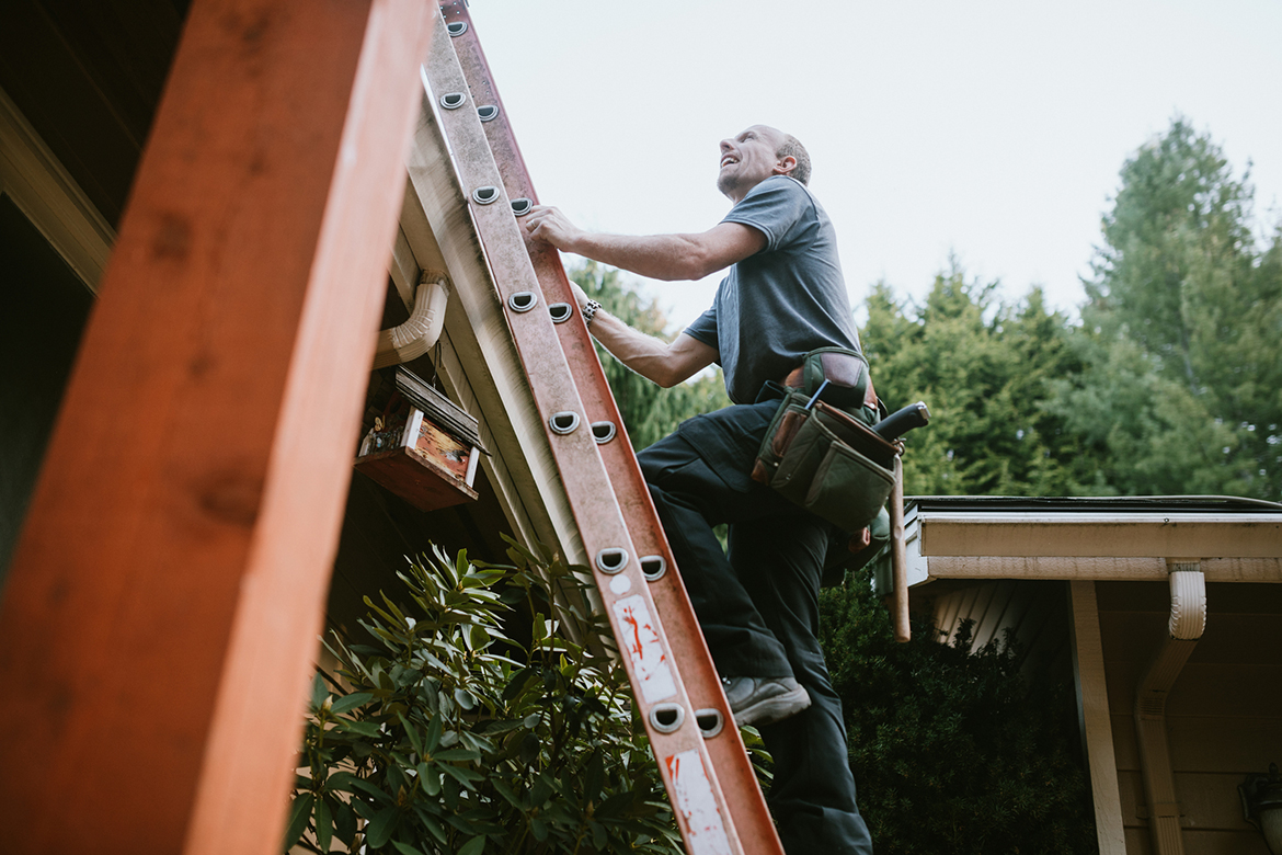 Man wearing tool belt climbs ladder onto roof