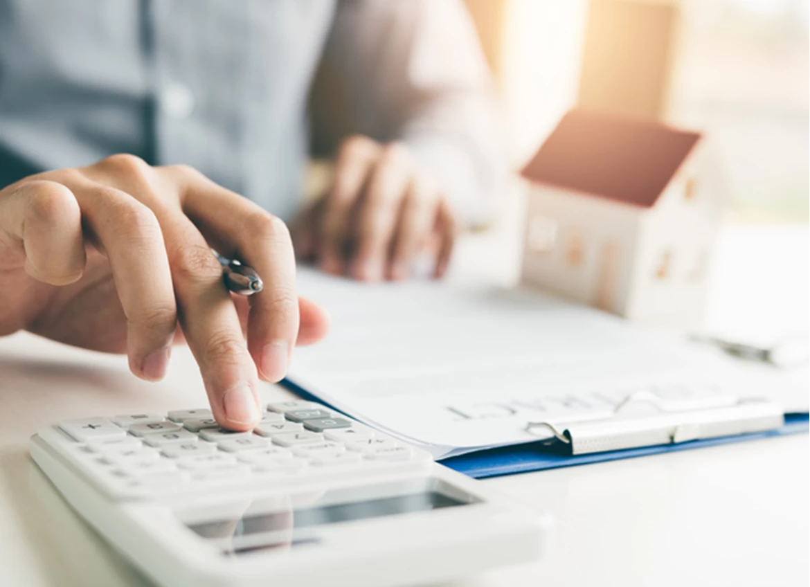 Person pushing buttons on calculator with clipboard and house figurine in background