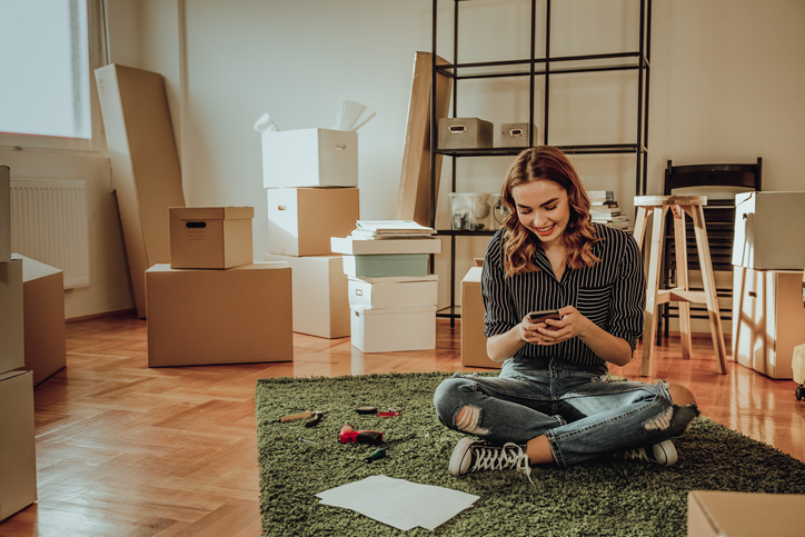 Woman sitting on floor looking at phone with boxes surrounding her