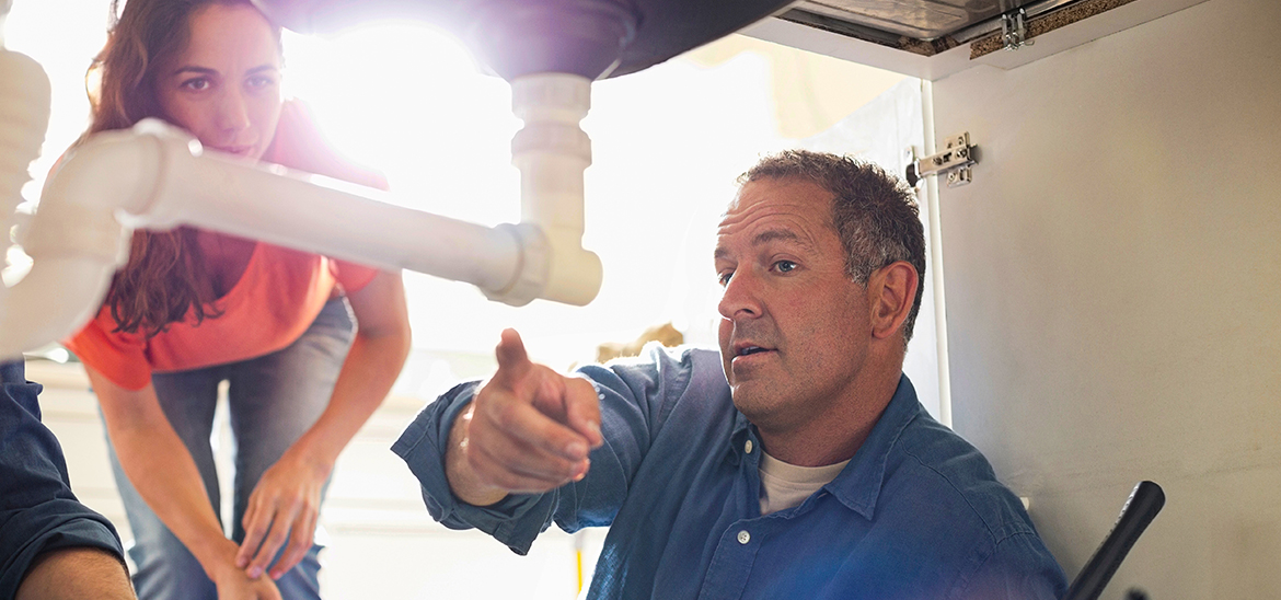 Repairman in blue pointing out a pipe to a woman leaning under a sink