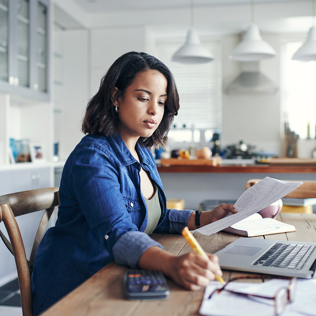 Woman sitting at a wood kitchen table running finances on a calculator and laptop