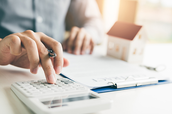 Person pushing buttons on calculator with clipboard and house figurine in background
